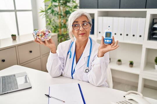 Middle age woman with grey hair wearing doctor uniform holding glucose monitor smiling looking to the side and staring away thinking.