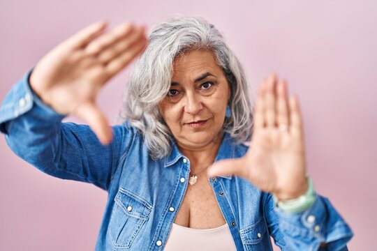 Middle age woman with grey hair standing over pink background doing frame using hands palms and fingers, camera perspective