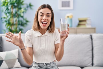 Hispanic woman drinking glass of water celebrating achievement with happy smile and winner expression with raised hand