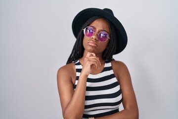 Young african american with braids wearing hat and sunglasses with hand on chin thinking about question, pensive expression. smiling with thoughtful face. doubt concept.