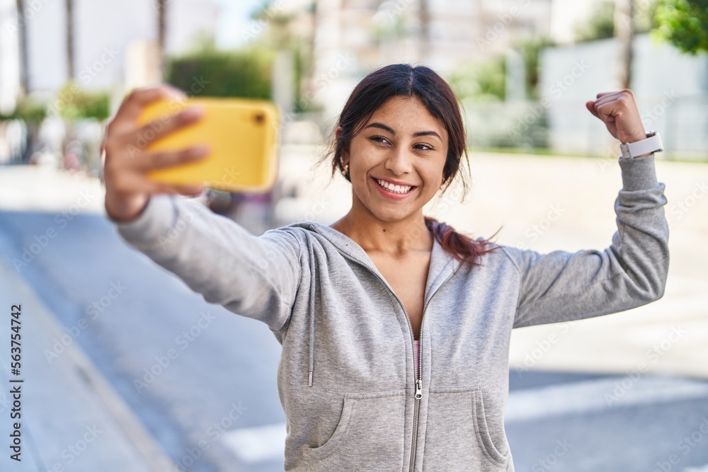 Poster Young hispanic woman make selfie doing strong gesture at street