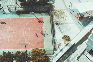 Aerial top view of a football/soccer and basketball fenced field, bordered by several trees, with two people training there, on a local school around Dubai