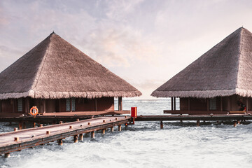A caption of two charming wooden bungalows and a wood walkway surrounded by crystal seawater in a dream-like environment in the Maldives island, in the background a lilac pastel cloudy sky
