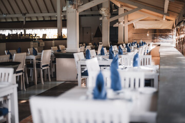 A capture using shallow depth of field and selective focus on the background of a restaurant that serves a resort, the white wooden dinner tables, already have silverware and blue napkins