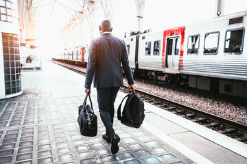 A backshot of a bald black elegant man well-dressed in a dark full-body suit, carrying a black backpack and a leather weekender, walking towards his train on the platform of the rail station - obrazy, fototapety, plakaty