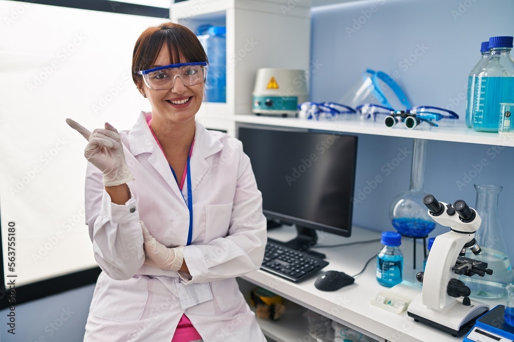 Sticker Young brunette woman working at scientist laboratory with a big smile on face, pointing with hand and finger to the side looking at the camera.