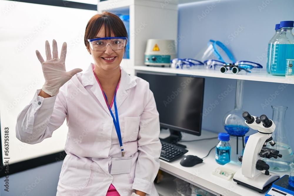 Poster young brunette woman working at scientist laboratory showing and pointing up with fingers number fiv