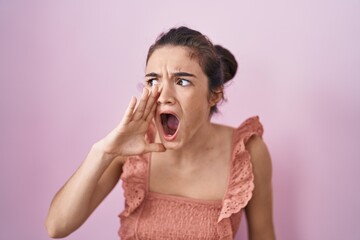 Young teenager girl standing over pink background shouting and screaming loud to side with hand on mouth. communication concept.