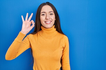 Young brunette woman standing over blue background smiling positive doing ok sign with hand and fingers. successful expression.