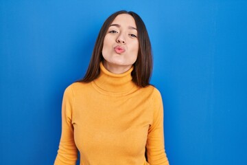 Young brunette woman standing over blue background looking at the camera blowing a kiss on air being lovely and sexy. love expression.