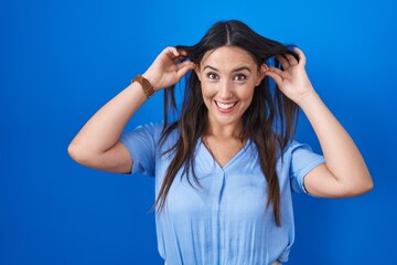 Young brunette woman standing over blue background smiling pulling ears with fingers, funny gesture. audition problem