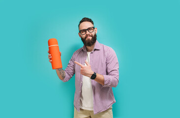 Young bearded charismatic hipster guy advertises, presents a thermos on a blue studio background