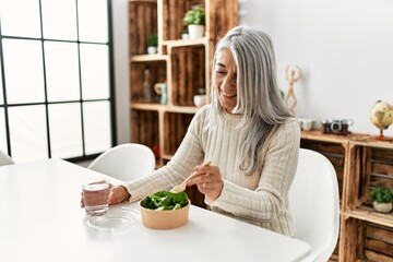 Middle age grey-haired woman eating salad sitting on table at home