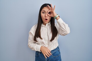 Young latin woman standing over blue background doing ok gesture shocked with surprised face, eye looking through fingers. unbelieving expression.