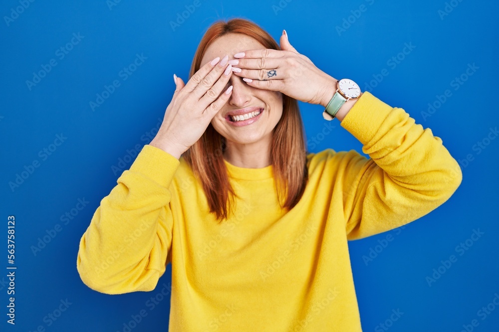 Wall mural Young woman standing over blue background covering eyes with hands smiling cheerful and funny. blind concept.