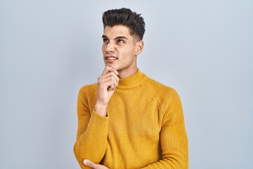 Young hispanic man standing over blue background thinking worried about a question, concerned and nervous with hand on chin