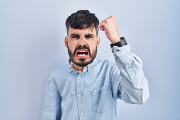 Young hispanic man with beard standing over blue background angry and mad raising fist frustrated and furious while shouting with anger. rage and aggressive concept.