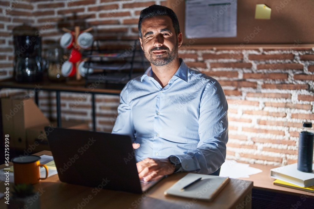 Canvas Prints Hispanic man with beard working at the office at night winking looking at the camera with sexy expression, cheerful and happy face.