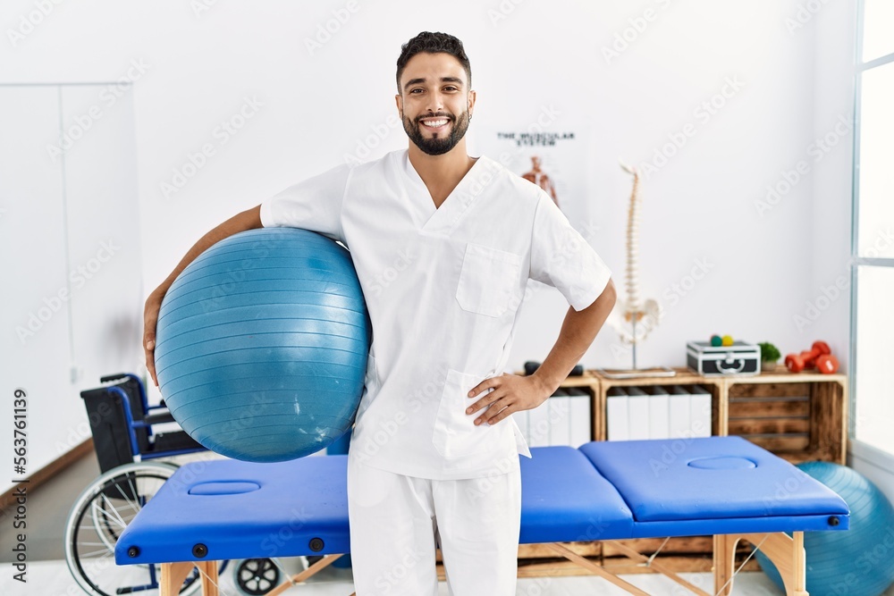 Wall mural young arab man wearing physiotherapist uniform holding fit ball at clinic