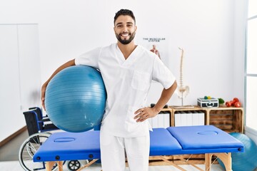 Young arab man wearing physiotherapist uniform holding fit ball at clinic - obrazy, fototapety, plakaty