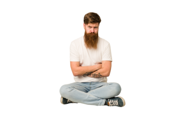 Young adult redhead with a long beard sitting on the floor isolated unhappy looking in camera with sarcastic expression.
