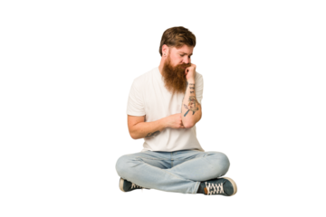Young adult redhead with a long beard sitting on the floor isolated who feels sad and pensive, looking at copy space.