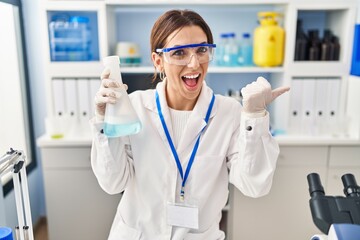 Young brunette woman working at scientist laboratory pointing thumb up to the side smiling happy...