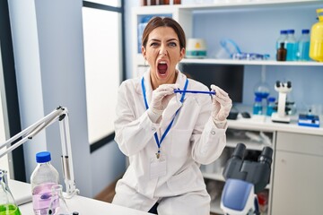 Young brunette woman working at scientist laboratory wearing safety glasses angry and mad screaming frustrated and furious, shouting with anger. rage and aggressive concept.
