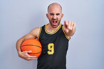 Young bald man with beard wearing basketball uniform holding ball pointing displeased and frustrated to the camera, angry and furious with you