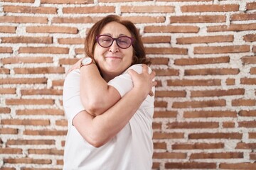 Senior woman with glasses standing over bricks wall hugging oneself happy and positive, smiling confident. self love and self care