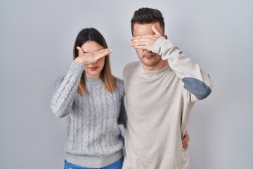 Young hispanic couple standing over white background covering eyes with hand, looking serious and sad. sightless, hiding and rejection concept