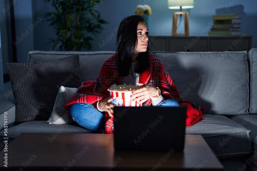 Canvas Prints Hispanic woman eating popcorn watching a movie on the sofa looking to side, relax profile pose with natural face and confident smile.