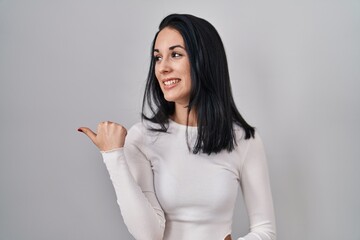 Hispanic woman standing over isolated background smiling with happy face looking and pointing to the side with thumb up.