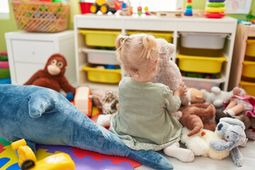 Adorable blonde girl playing with teddy bear sitting on floor at kindergarten