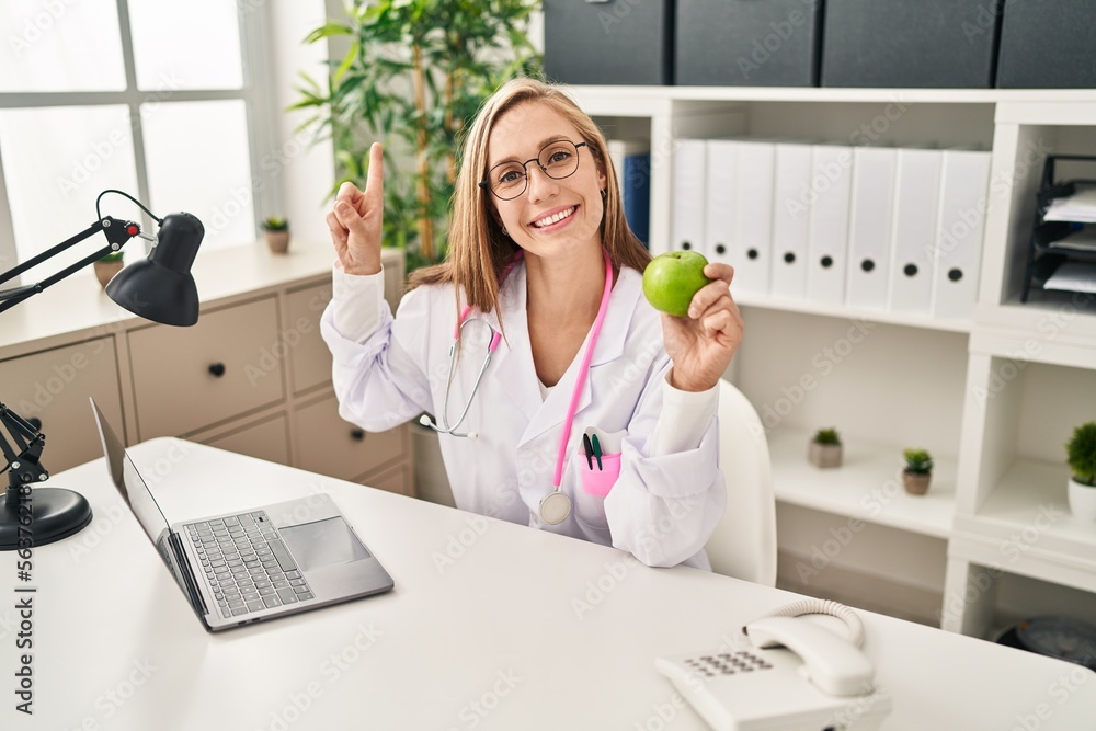 Poster young blonde doctor woman holding green apple at the clinic smiling happy pointing with hand and fin