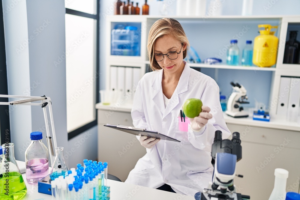 Sticker young blonde woman scientist holding apple reading report at laboratory