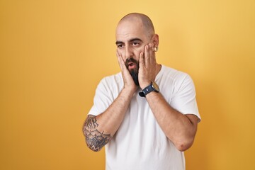 Young hispanic man with beard and tattoos standing over yellow background tired hands covering face, depression and sadness, upset and irritated for problem