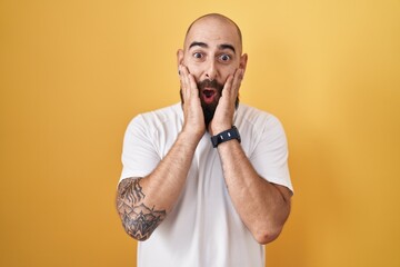 Young hispanic man with beard and tattoos standing over yellow background afraid and shocked, surprise and amazed expression with hands on face