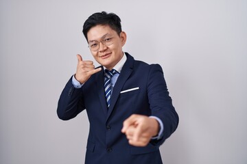 Young asian man wearing business suit and tie smiling doing talking on the telephone gesture and pointing to you. call me.