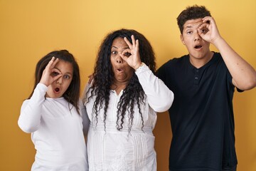 Family of mother, daughter and son standing over yellow background doing ok gesture shocked with surprised face, eye looking through fingers. unbelieving expression.