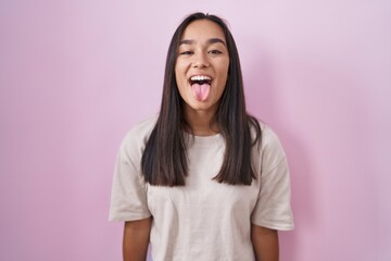 Young hispanic woman standing over pink background sticking tongue out happy with funny expression....