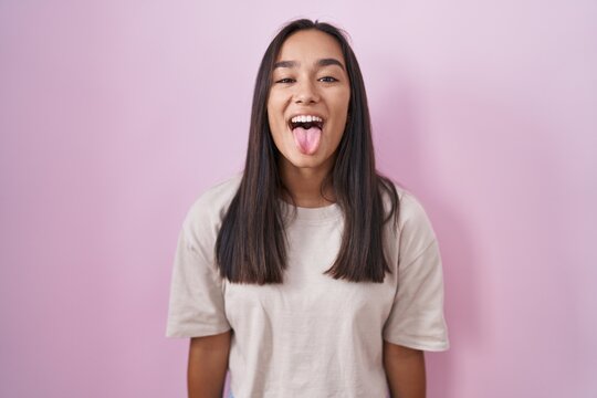 Young hispanic woman standing over pink background sticking tongue out happy with funny expression. emotion concept.
