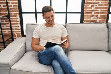 Young hispanic man reading book sitting on sofa at home