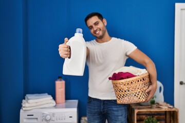 Young hispanic man holding basket with clothes and detergent bottle at laundry room
