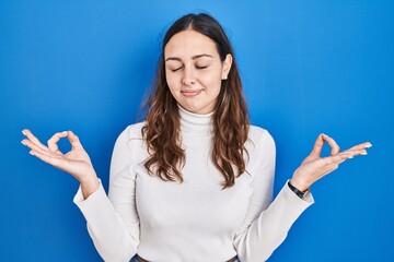 Young hispanic woman standing over blue background relaxed and smiling with eyes closed doing meditation gesture with fingers. yoga concept.