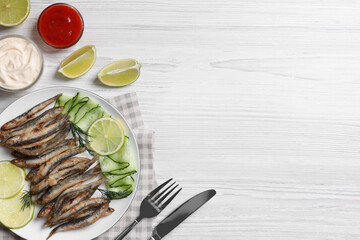 Delicious fried anchovies with lime, cucumber and sauces served on white wooden table, flat lay. Space for text