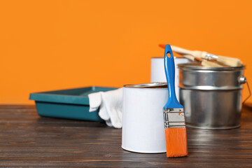 Can with paint, brush and renovation equipment on wooden table against orange background