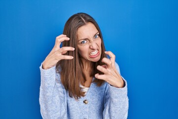 Young woman standing over blue background shouting frustrated with rage, hands trying to strangle, yelling mad