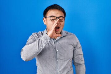 Obraz premium Young chinese man standing over blue background shouting and screaming loud to side with hand on mouth. communication concept.