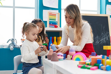 Naklejka premium Teacher with girls playing with geometry blocks sitting on table at kindergarten